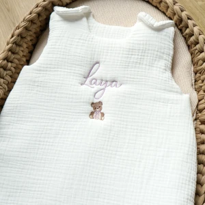 Gigoteuse bébé blanche en gaze de coton, brodée avec le prénom 'Laya' en fil violet et un petit ours brun en dessous, posée dans un couffin en osier.